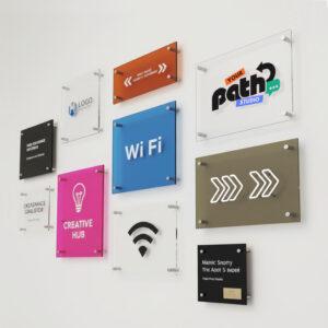 Wall display of bespoke acrylic signs featuring custom logos, directional signage, WiFi symbols and branded panels, mounted with standoff fixings in a modern office environment.