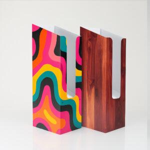 Two free-standing magazine and brochure dispensers placed side by side on a white surface. The left dispenser features a bright, retro-style abstract pattern in pink, orange, teal, yellow and black, while the right dispenser has a realistic dark wood-grain finish. Both units have a tall vertical front opening that reveals the white interior.