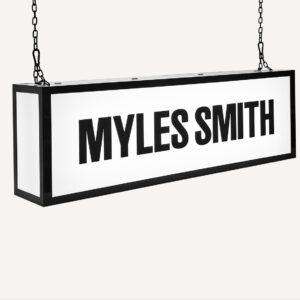A ceiling hanging illuminated sign with "Myles Smith" on the front.