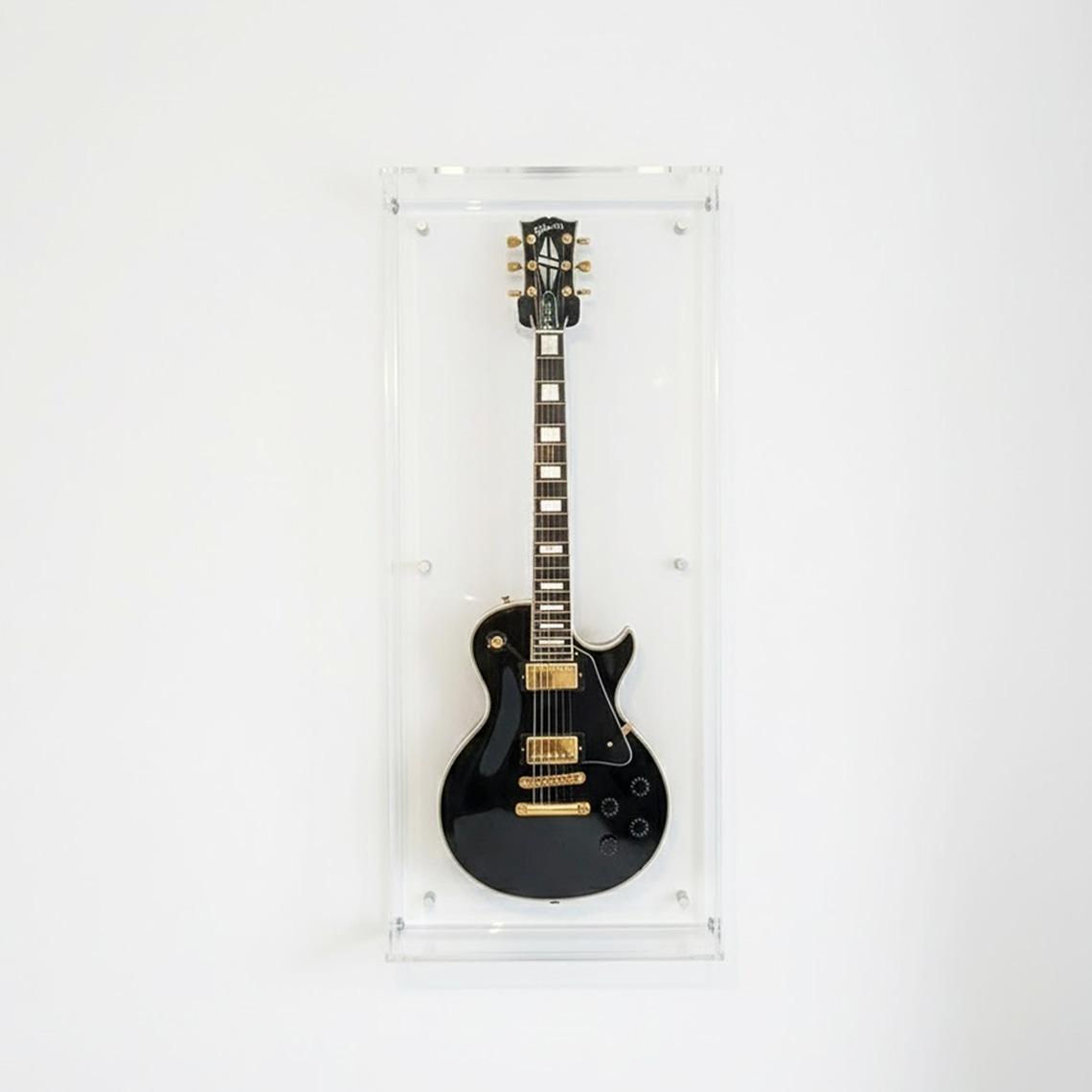 Front-facing view of a wall-mounted clear acrylic guitar display case showcasing an electric guitar on a white wall.