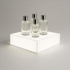 A minimalist countertop illuminated plinth displays four elegant perfume bottles in clear and frosted glass, arranged neatly on a reflective surface against a neutral background.