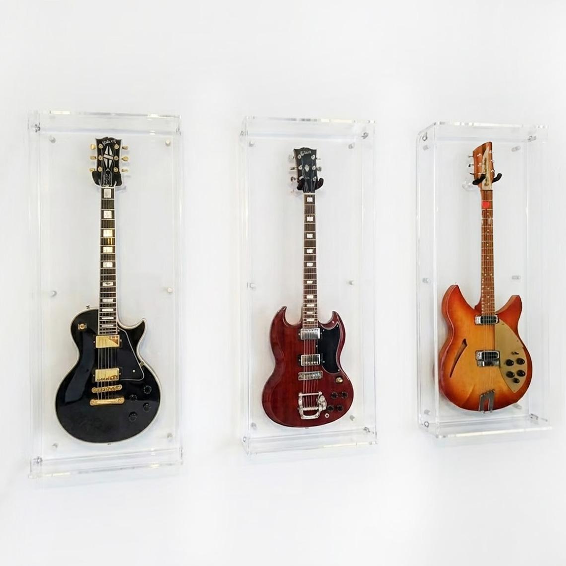 Three wall-mounted clear acrylic guitar display cases holding electric guitars, arranged side by side on a white wall.