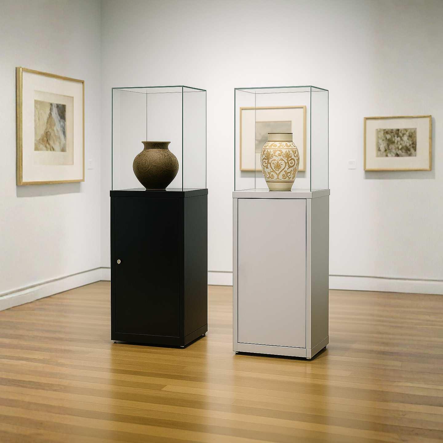 A lockable black glass display plinth and a silver glass display case plinth in a gallery with vases on display inside of the cases.