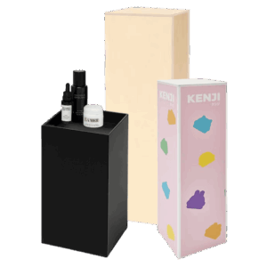 Three modern display plinths of different styles and heights arranged together, including a black acrylic plinth holding cosmetic products, a natural plywood plinth, and a pastel pink branded plinth with colorful graphics.