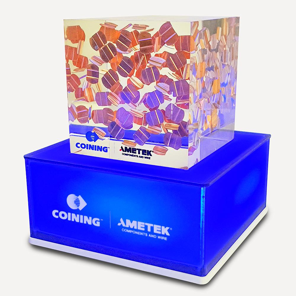 Blue illuminated pedestal base with an "Ametek logo" on the front and a decorative patterned transparent block on top.