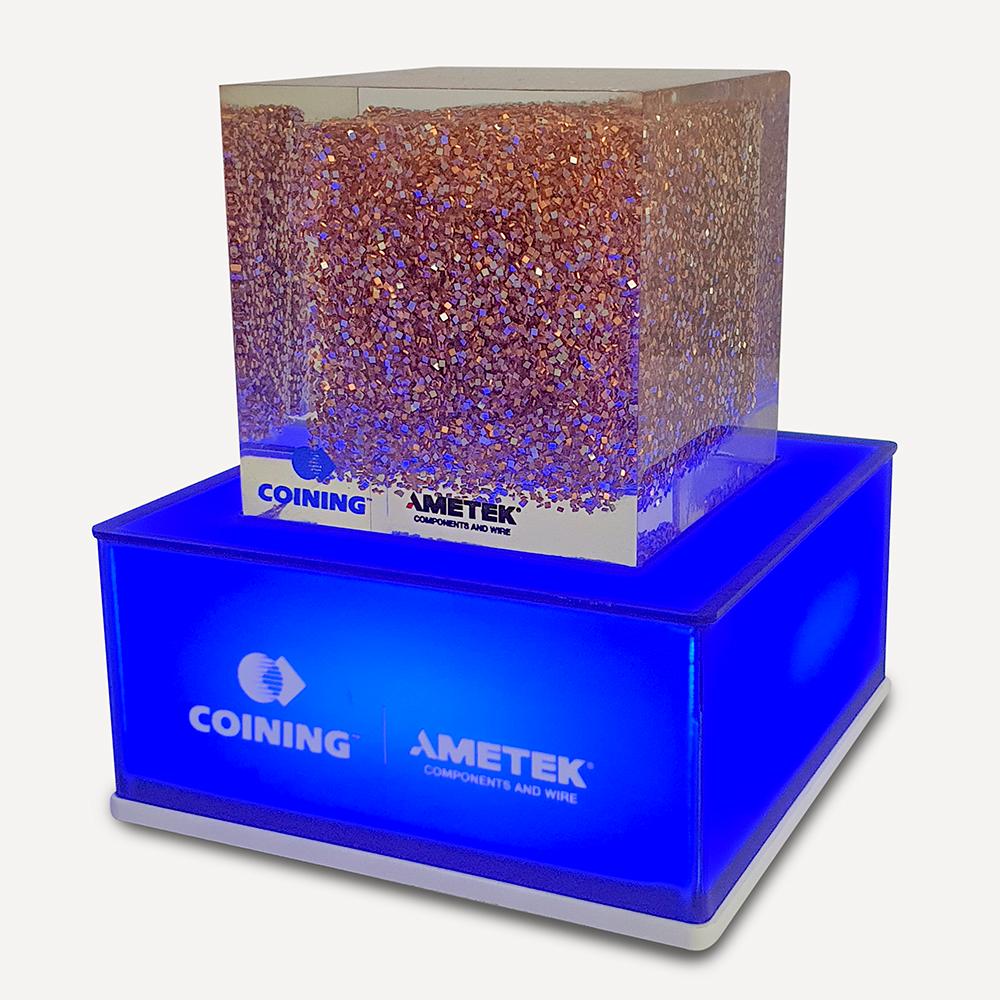 A blue illuminated pedestal base with an "Ametek logo" on the front and a decorative patterned transparent block on top.