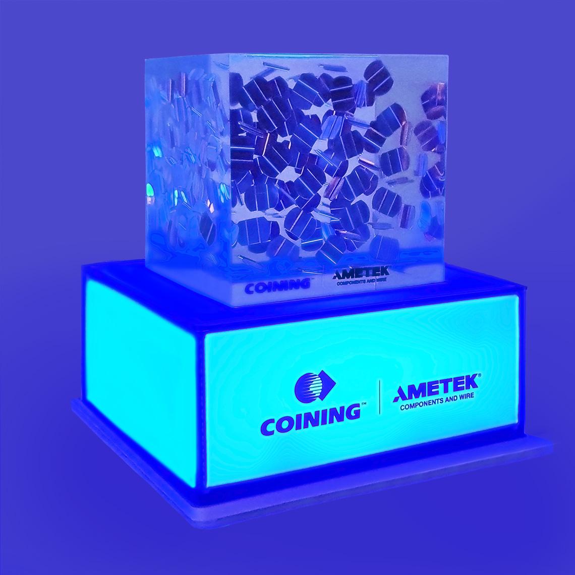 A fluorescent blue illuminated pedestal base with an "Ametek logo" on the front and a decorative patterned transparent block on top with a blue background.