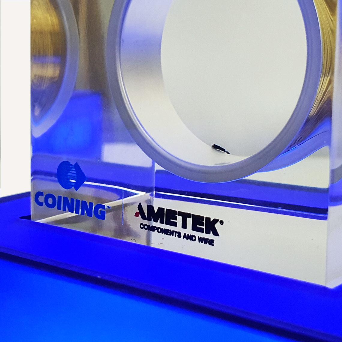 A close up of a blue illuminated pedestal base and a clear acrylic block on top with an "Ametek logo" on the front.