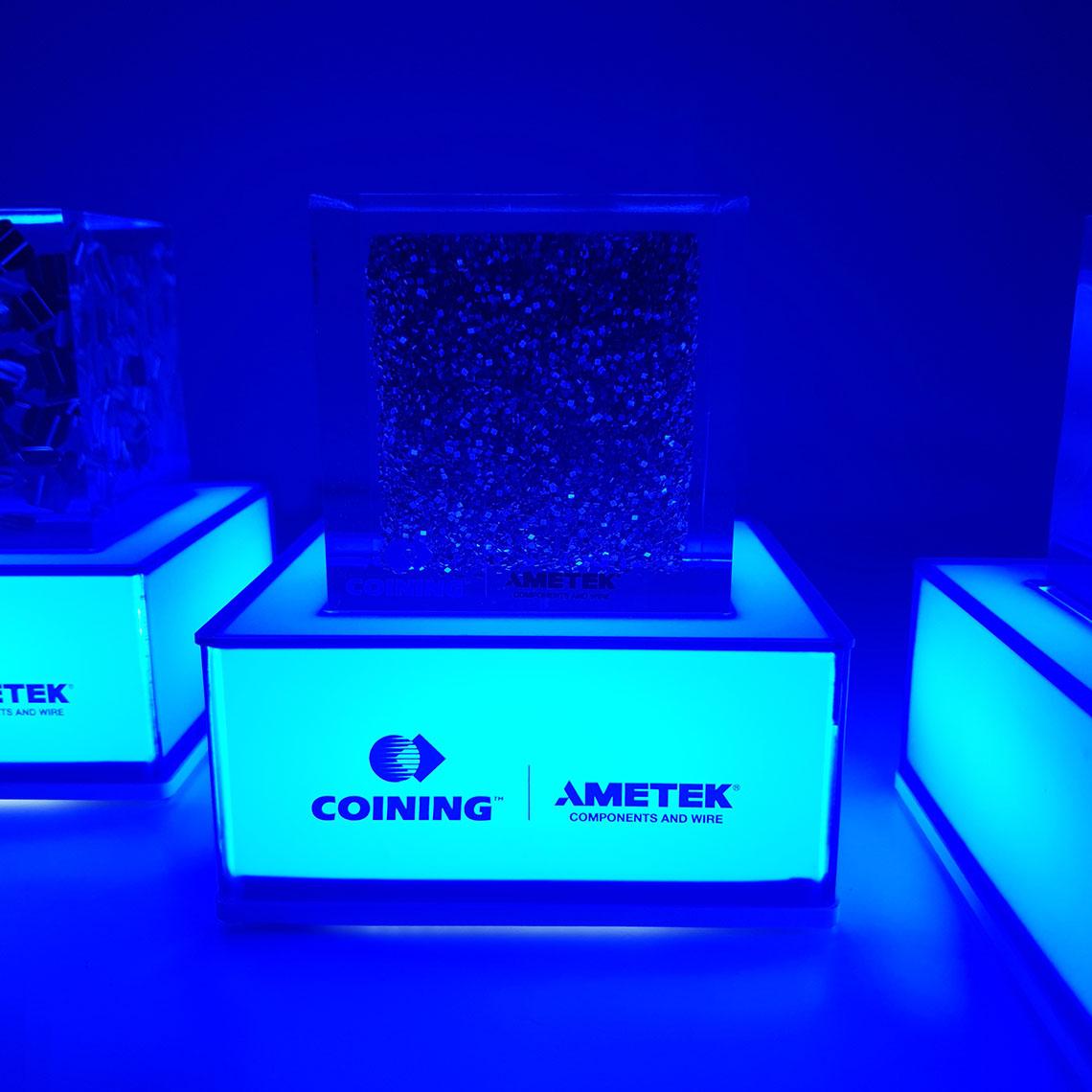 A set of fluorescent blue illuminated pedestal base with an "Ametek logo" on the front and a decorative patterned transparent block on top with a blue background.