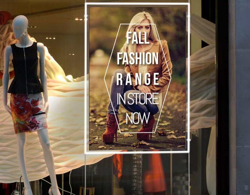 A ceiling hanging digital advertising screen in a shop window with an autumn clothing ad on the screen.