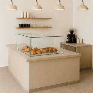 Large L-shaped glass sneeze guard on a beige café countertop displaying fresh croissants and bread loaves in a modern minimalist bakery setting.