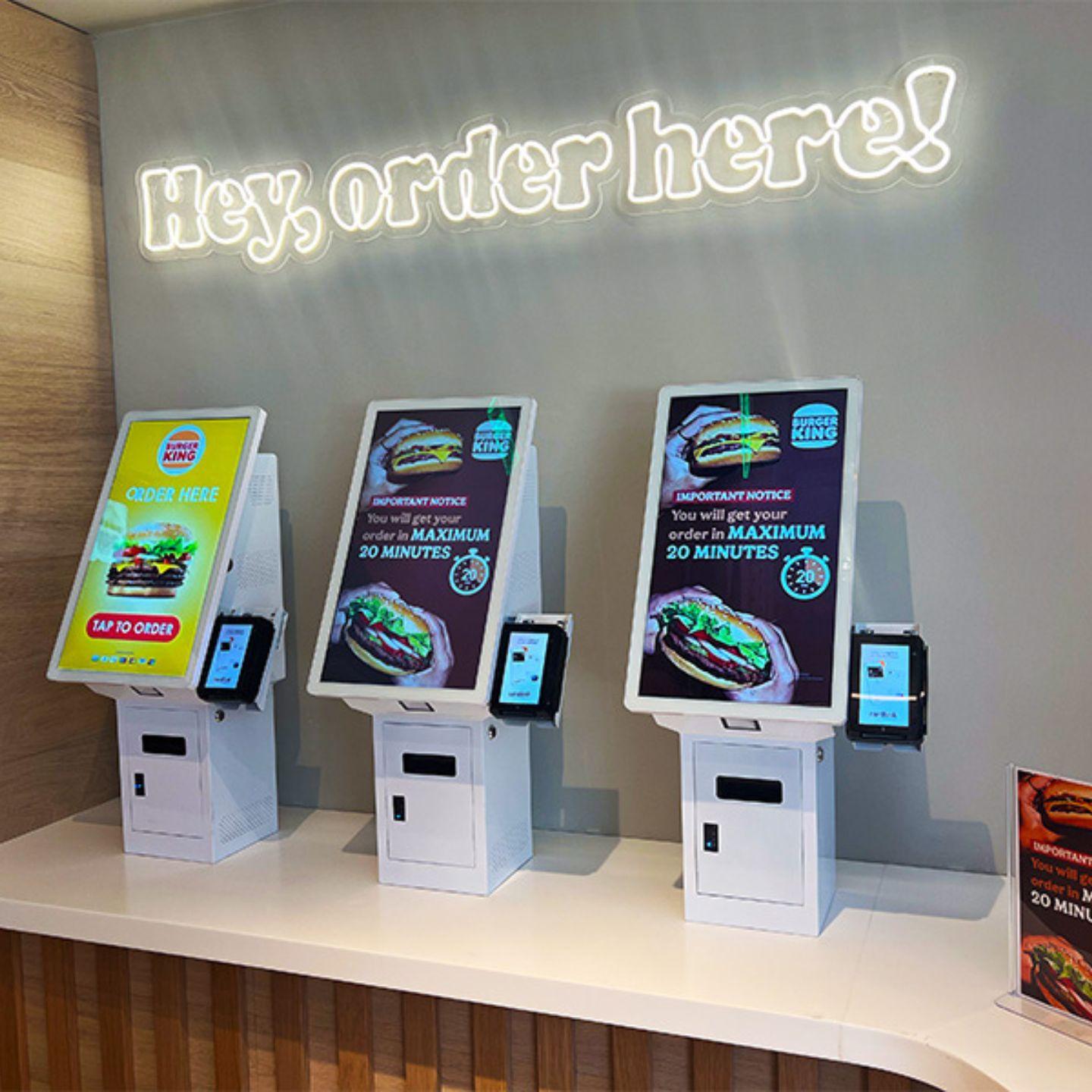 Fast-food touch screen checkout kiosks—three countertop self-service ordering stations with card readers under ‘Hey, order here!’ sign.
