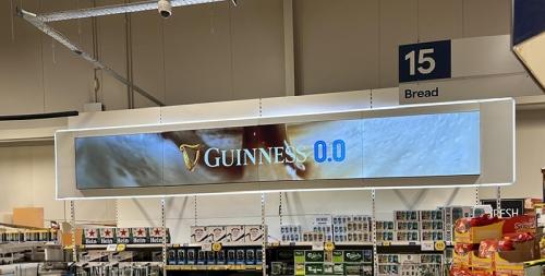 Supermarket aisle with an ultra-wide digital banner above shelves displaying a Guinness 0.0 advertisement under aisle sign ‘15 Bread’.
