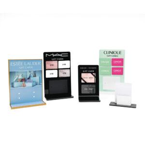 A range of custom branded countertop gift card displays, including designs for Estée Lauder, MAC, Bobbi Brown, and Clinique. The displays feature different colours, styles, and sizes, each showcasing gift cards on acrylic stands.