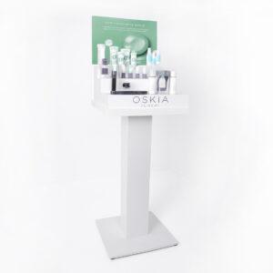 White freestanding display plinth showcasing OSKIA London skincare products, arranged on a stepped shelf with a green informational backdrop, photographed in a clean white studio setting.