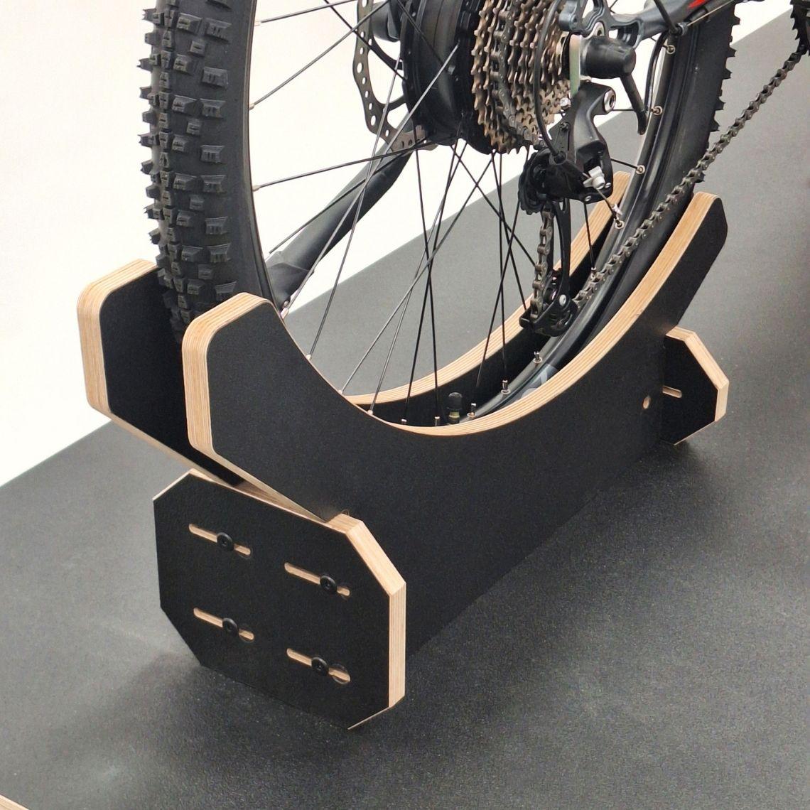 Close-up of the adjustable plywood rear wheel support on the bike display podium, showing curved black melamine panels and slidable mounting slots.