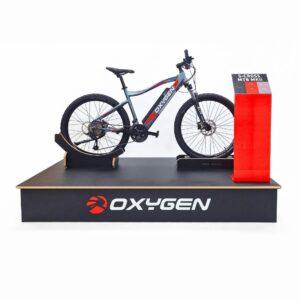 Front-facing view of a custom-branded Oxygen bike display podium featuring an S-Cross MTB MKII on a plywood stand with adjustable supports and a red magnetic POS graphic panel.