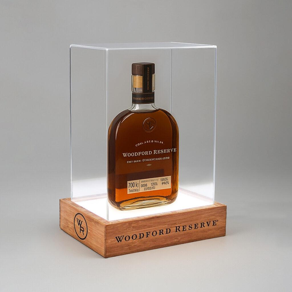 An illuminated bottle display case featuring a Woodford Reserve whiskey bottle showcased on a glowing base with a wooden plinth and clear acrylic cover, designed for premium retail and branded product presentation.