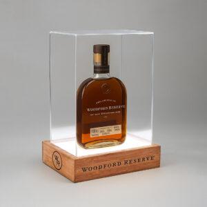 An illuminated bottle display case featuring a Woodford Reserve whiskey bottle showcased on a glowing base with a wooden plinth and clear acrylic cover, designed for premium retail and branded product presentation.