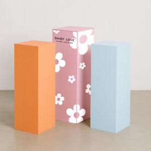 Three pastel-colored acrylic display plinths—one orange, one pink, and one light blue—stand side by side on a smooth, neutral floor against a plain white backdrop. Each plinth has clean, sharp edges and a matte finish, arranged in varying heights for a modern, minimalist presentation. The pink plinth features Marc Jacobs Daisy Love Branding.
