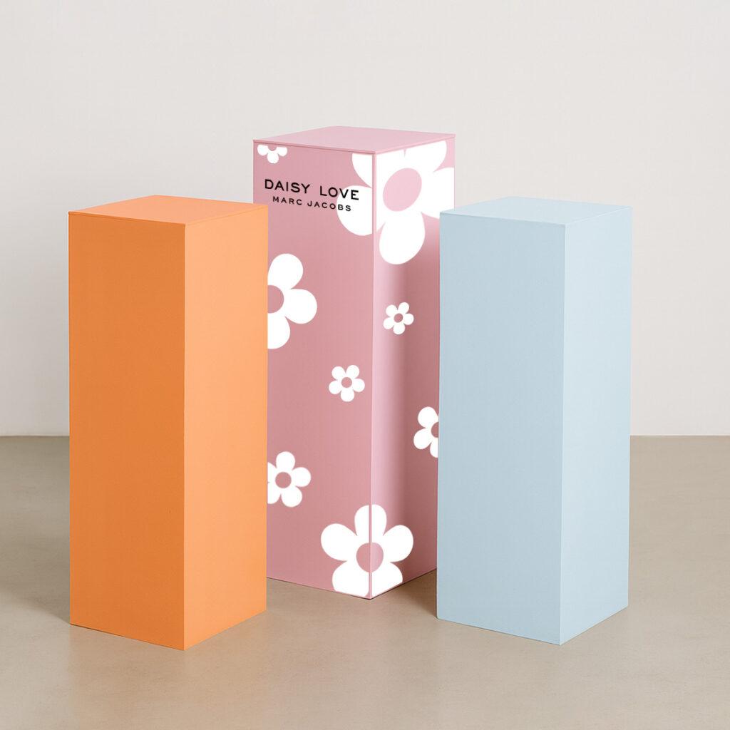 Three pastel-colored acrylic display plinths—one orange, one pink, and one light blue—stand side by side on a smooth, neutral floor against a plain white backdrop. Each plinth has clean, sharp edges and a matte finish, arranged in varying heights for a modern, minimalist presentation. The pink plinth features Marc Jacobs Daisy Love Branding.