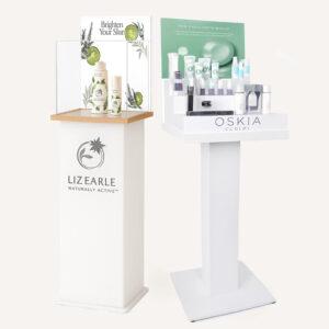 Two branded capped display plinths showcasing Liz Earle and Oskia skincare products, featuring a clear display case and elevated retail presentation.