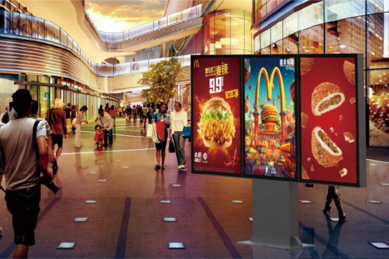Three digital advertising screens with mcdonalds promotions on, in a shopping mall with people walking around.