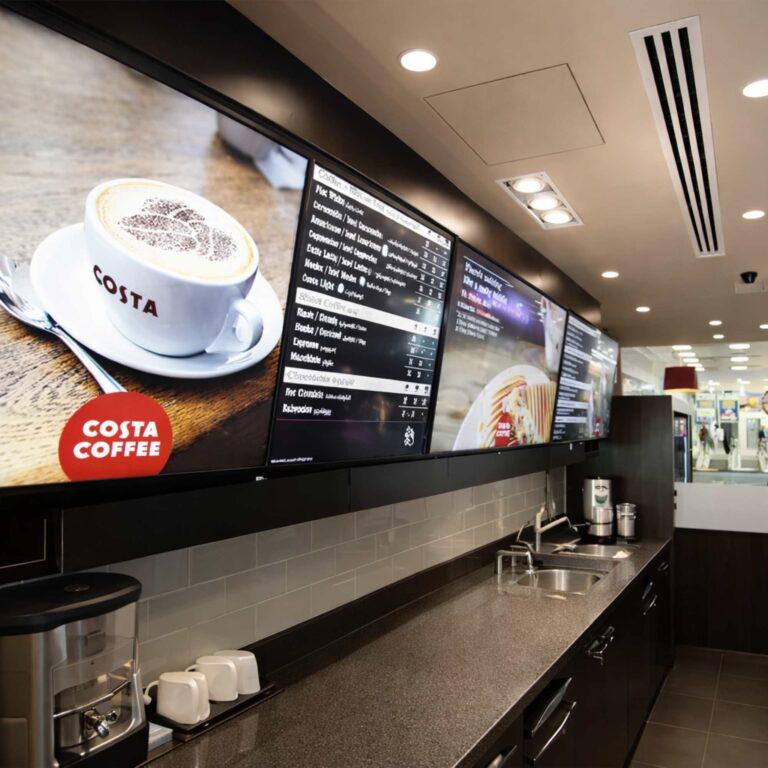Four ceiling mounted digital screens with costa advertisements on them in a cafe.