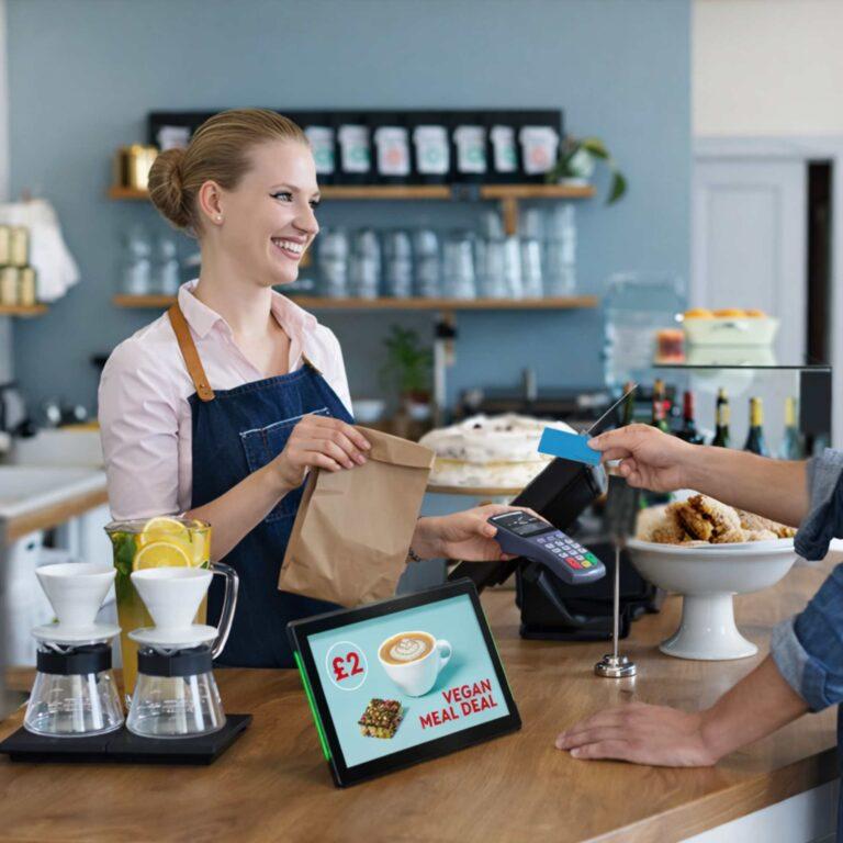 A cafe worker smiling, handing a bag of products over to someone reaching to pay with their card. There is a countertop digital screen advertising promotions.
