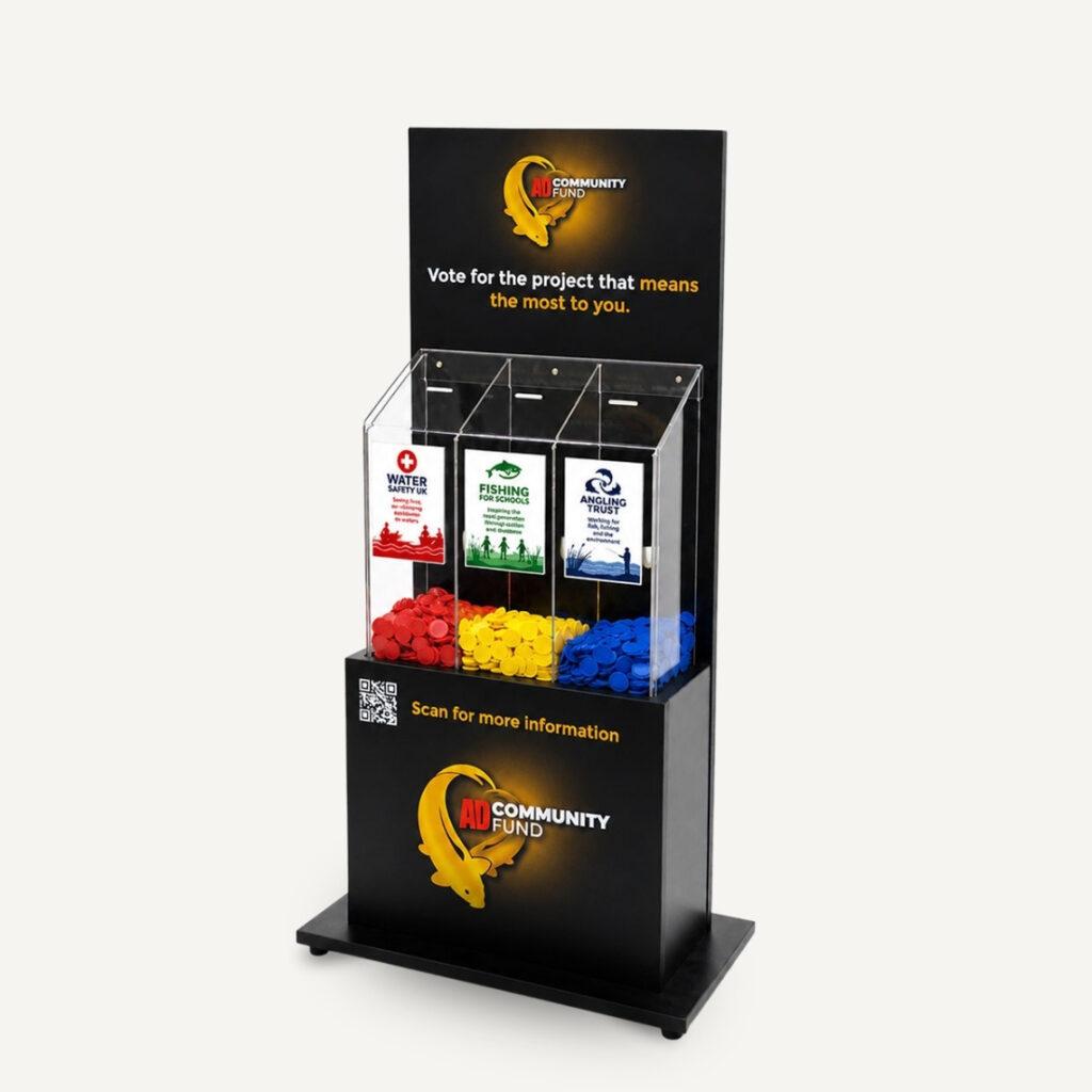 Angled view of a black AD Community Fund voting stand with three transparent ballot compartments containing red, yellow, and blue voting tokens. Each section is labelled with a different community initiative — Water Safety UK, Fishing for Schools, and Angling Trust — encouraging visitors to vote for the project they support most. A QR code on the front provides access to more information.