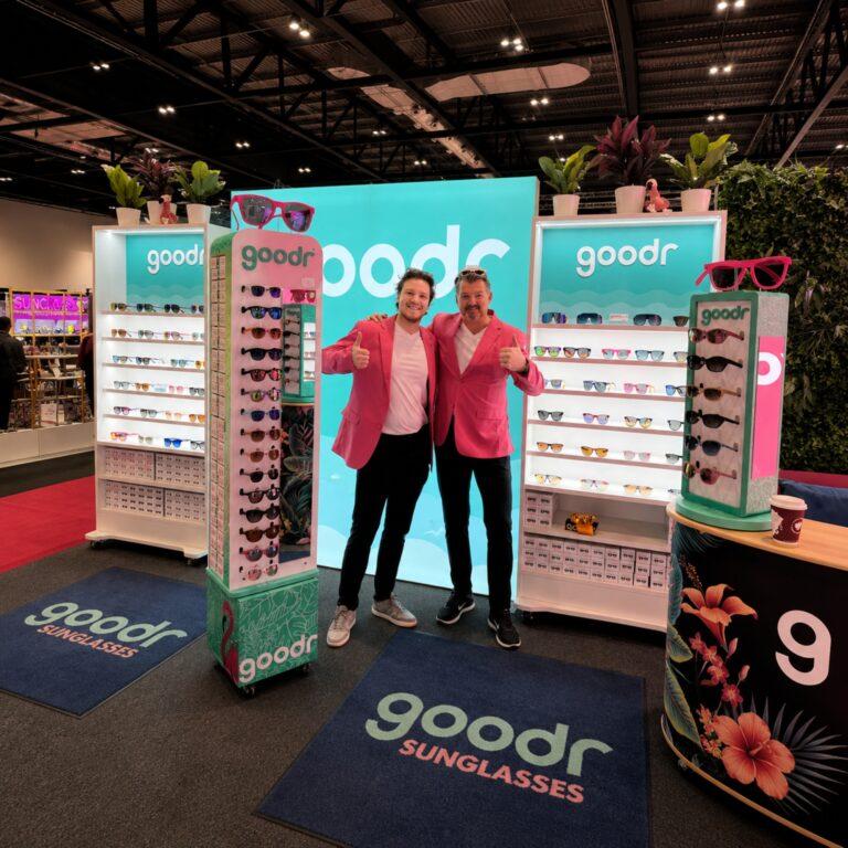 Two brand ambassadors smiling with their thumbs up, stood amongst some floorstanding modular sunglasses displays including illuminated shelving, sunglasses display with castor wheels and a countertop display.