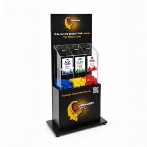 Freestanding AD Community Fund voting display with three clear collection compartments filled with blue, yellow, and red voting counters. The compartments are labelled for Angling Trust, Fishing for Schools, and Water Safety UK, inviting visitors to vote for the project they value most. The black branded stand includes campaign messaging and a QR code for further information.
