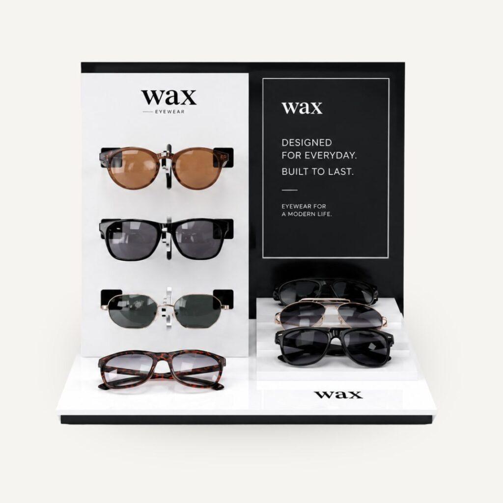 Front view of the bespoke WAX eyewear countertop display manufactured from black and white acrylic, featuring three sunglasses hooks, a three-tier display platform, and interchangeable branded graphic panel.